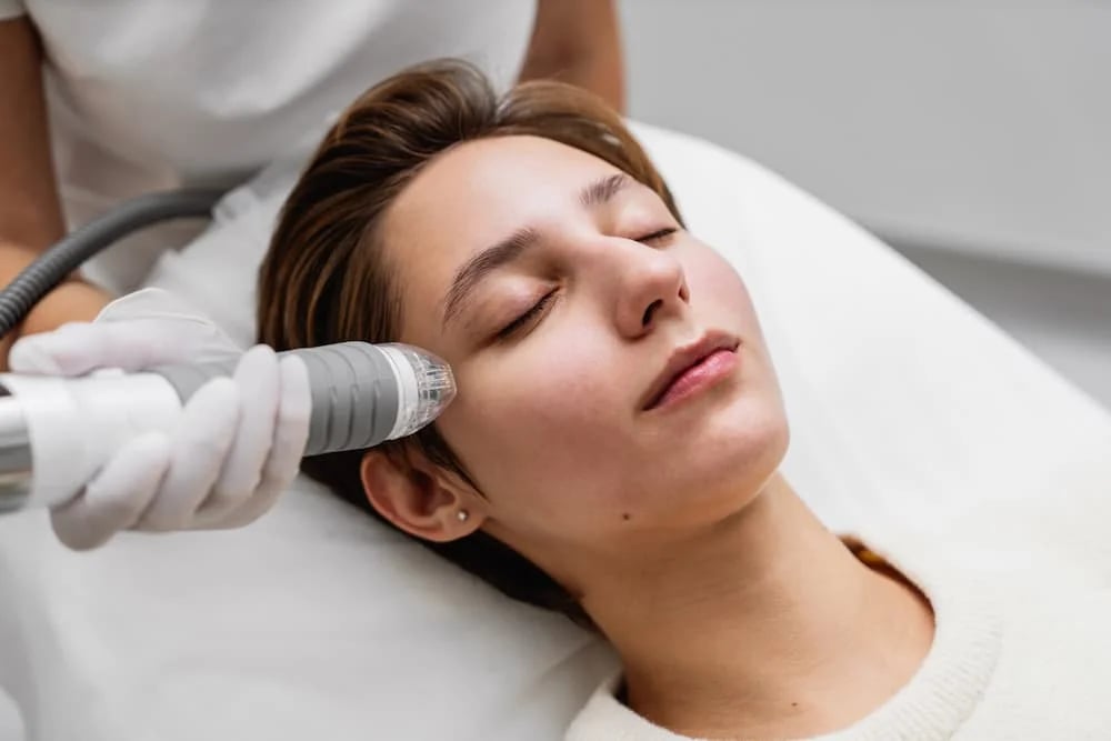 A patient undergoing an RF microneedling treatment. A medical professional in white gloves uses a specialized handheld device to deliver radiofrequency energy and micro-needles to the patient's cheek for skin tightening and collagen stimulation.