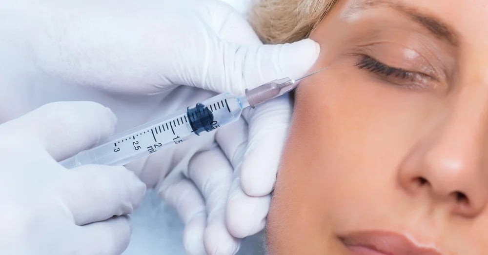 A close-up of a gloved hand administering a facial injection near the eye area of a woman with closed eyes against a light background.