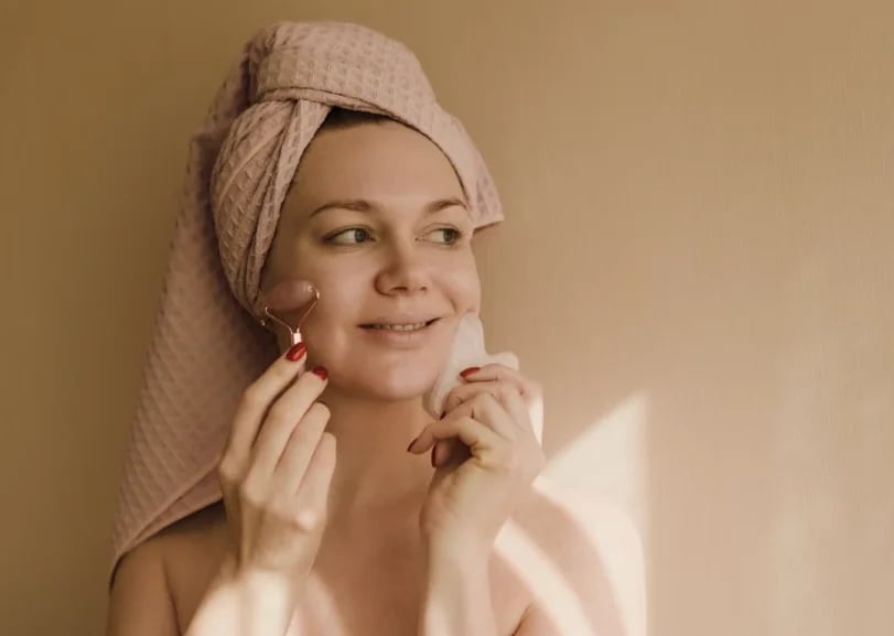 Woman in a towel wrap smiling while using a facial roller and gua sha stone