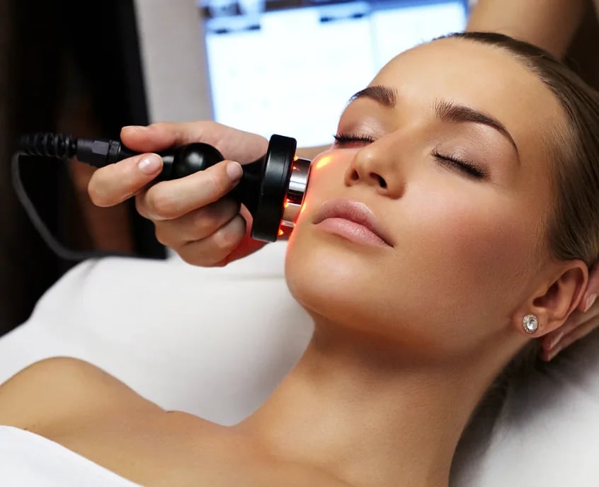Esthetician holding a handheld facial device against the cheek of a relaxed woman lying back with her eyes closed