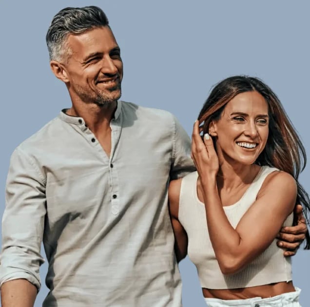 Smiling man and woman in light linen clothing standing together against a pale blue backdrop