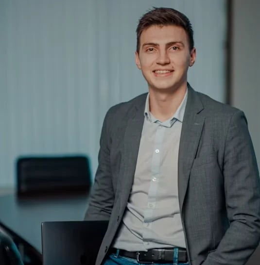 Smiling man in a gray blazer leaning against a desk in an office